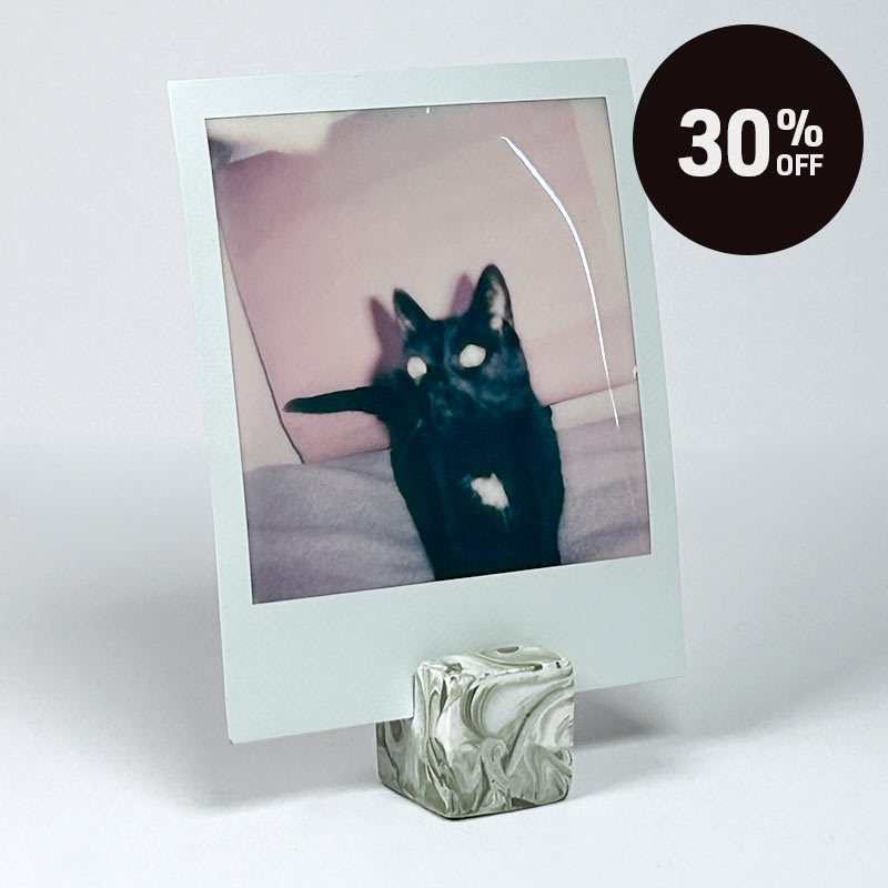 A square instant-film photo frame holder displaying a polaroid photo of a black cat, standing on a marbled cube base against a light grey background. A dark circular '30% OFF' sale badge appears in the top right corner.