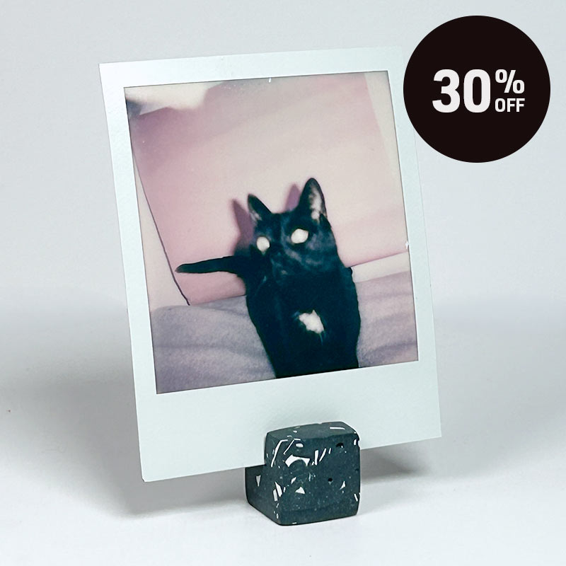 A Polaroid-style instant photo print on a small dark stand, featuring a black cat with a white chest patch. A dark brown circular '30% OFF' sale badge is overlaid in the top right on a light gray studio background.