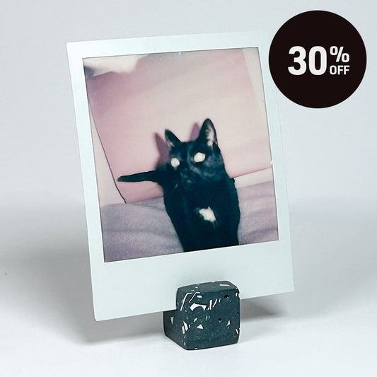 A Polaroid-style instant photo print on a small dark stand, featuring a black cat with a white chest patch. A dark brown circular '30% OFF' sale badge is overlaid in the top right on a light gray studio background.