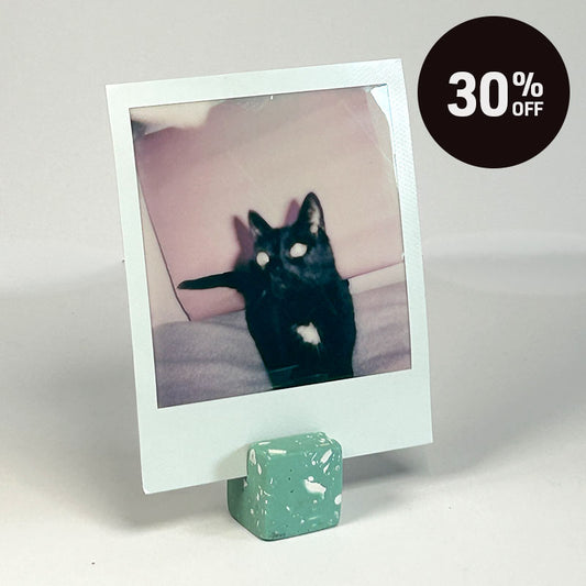 A square polaroid photo print stands on a small sage green terrazzo photo holder against a light grey background. The printed photo shows a black cat sitting on a pink cushion with a black circular '30% OFF' badge in the top right corner.