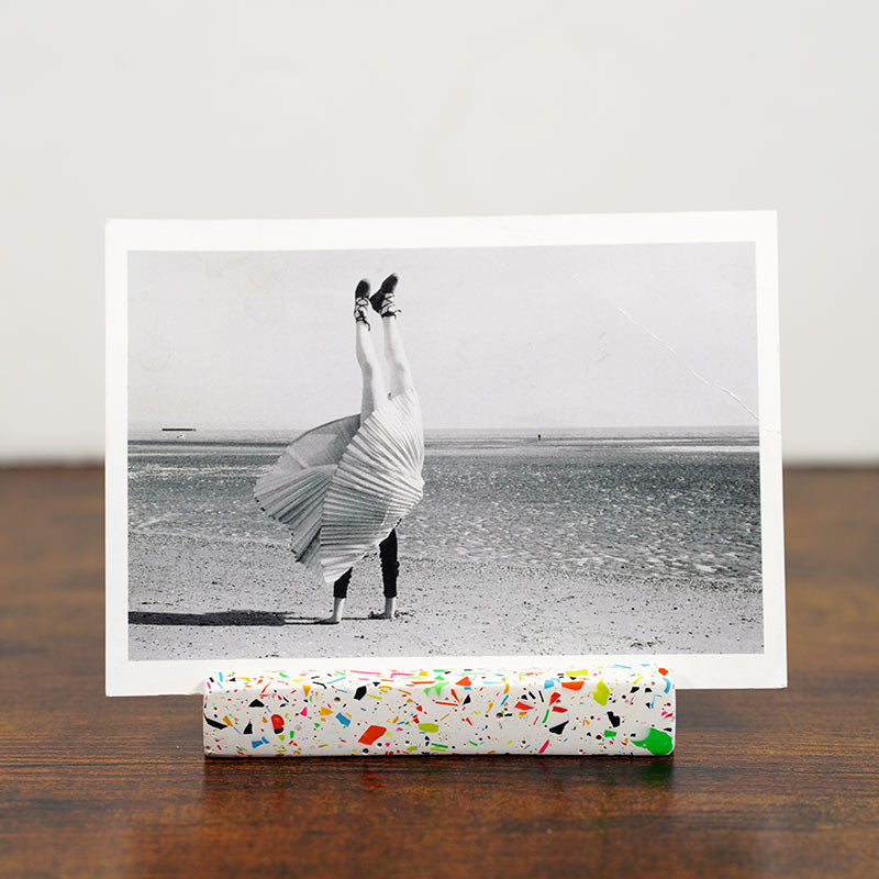 A black and white art print of a person dancing on a beach, displayed on a rainbow terrazzo photo holder on a wooden surface against a light grey wall.