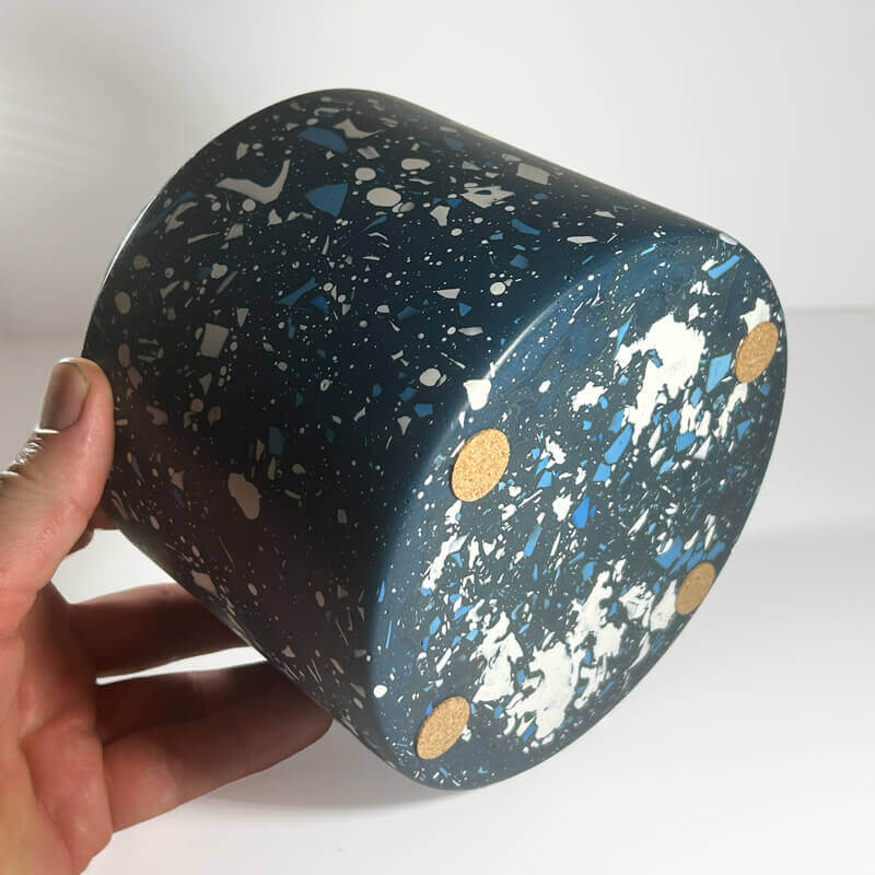 A hand holding a cylindrical planter pot with a dark blue terrazzo pattern featuring white, blue, and gold flecks, set against a plain white background.