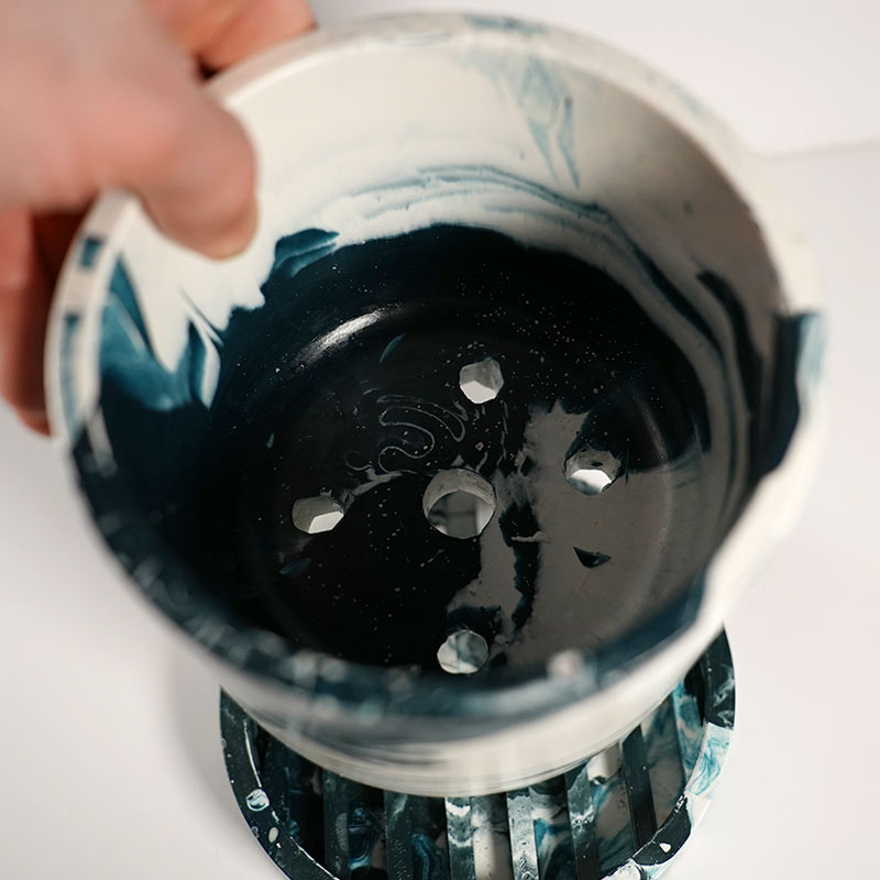 Close-up of a hand holding a navy marbled plant pot with drainage holes with a separate water tray on a white background