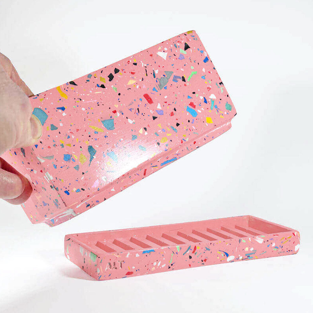 Hand holding a pink rectangular plant pot with colourful speckles terrazzo with drainage holes and a water tray on a white background