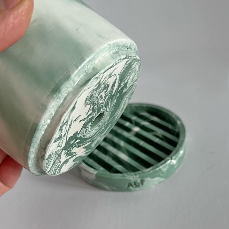 Moss Green and white marbled plant pot with 'ALP' branding with a water tray held by a hand on a gray background