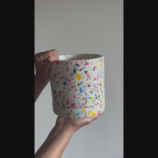 Hand holding a large rainbow terrazzo Jesmonite plant pot with drainage holes and matching water tray, a modern handmade indoor planter