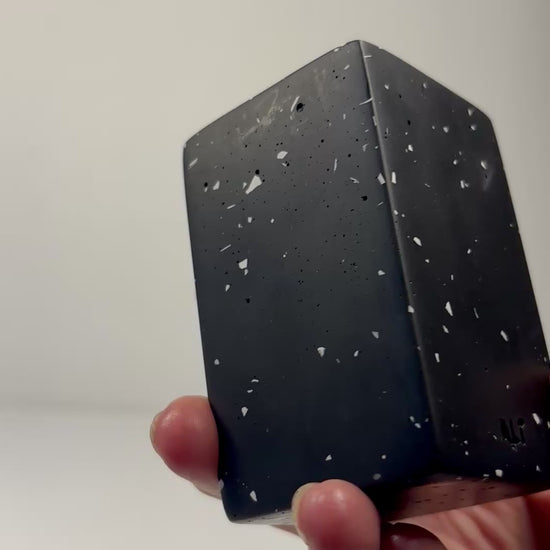 Hand holding a black with white terrazzo rectangular shaped tall tea light candle holder with a tea light candle inside on a plain background
