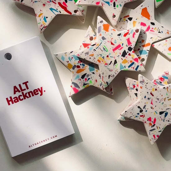 Rainbow-confetti star-shaped Christmas ornaments on a white surface, with a festive display card showing how to thread a gold string through.