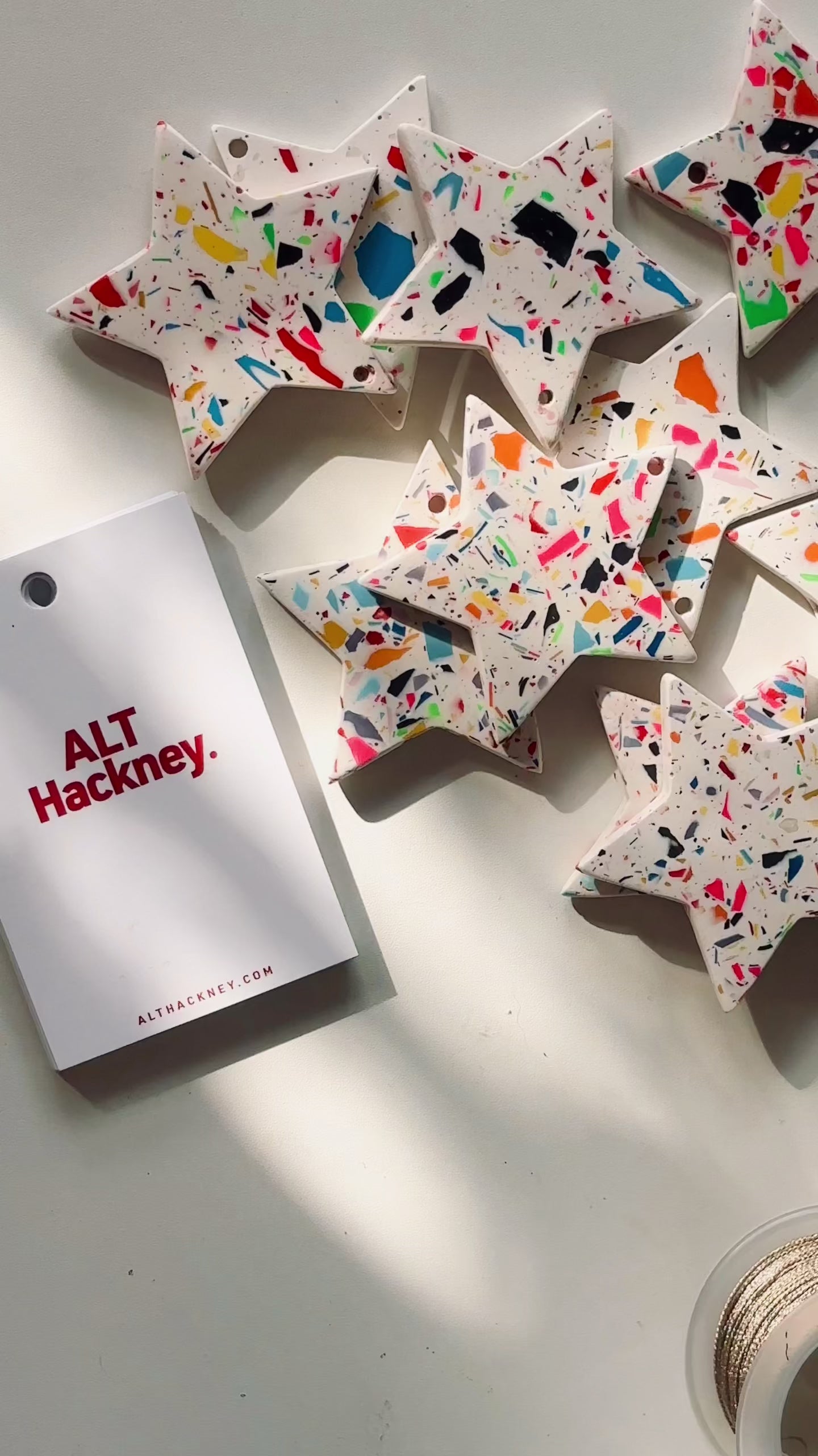Rainbow-confetti star-shaped Christmas ornaments on a white surface, with a festive display card showing how to thread a gold string through.