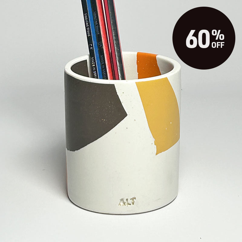 A white pencil holder with abstract geometric colour blocks in dark gray, mustard yellow, and orange, containing several pencils. A black circular '60% OFF' promotional sticker is in the top right corner.