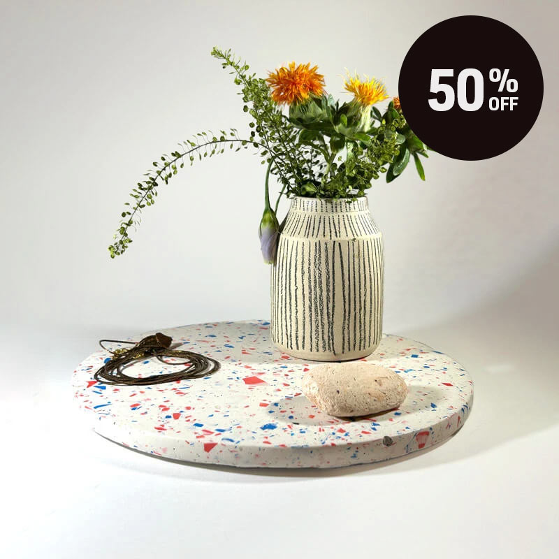 A round terrazzo tray with a white base and red and blue chips with a vase holding orange and yellow flowers, alongside a dark gold jewerlly and a small stone, against a plain light background. A black circular badge in the top right reads '50% OFF'.