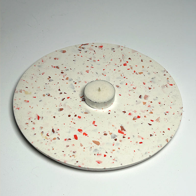 A round white concrete tea light holder with a terrazzo pattern of red, brown, and pink chips, featuring a central cylindrical opening for a tea light, shown on a neutral light grey background.