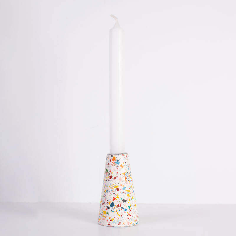 A tall white taper candle standing upright in a white and rainbow terrazzo patterned taper candle holder with a silver-toned top, shown on a white background