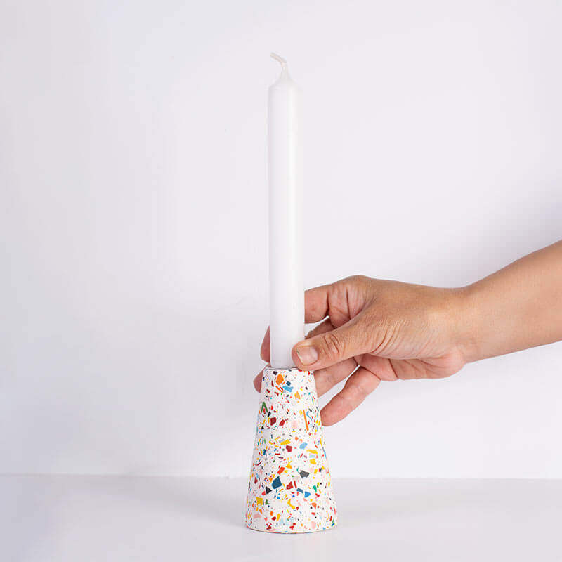 A hand holding a white taper candle in a white and rainbow terrazzo patterned taper candle holder with a silver-toned top, shown on a white background