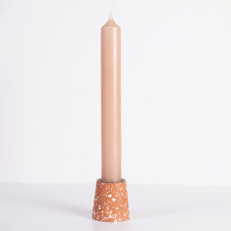 A slim beige taper candle standing upright in a terracotta-coloured white terrazzo taper candle holder against a plain white background.