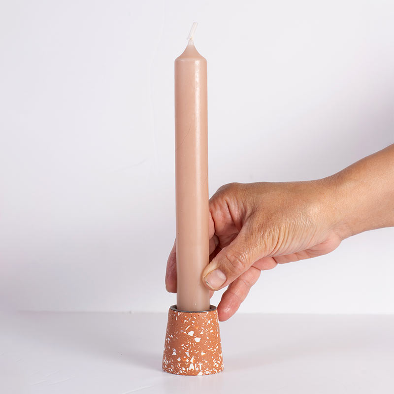 A hand holding a tall, slim beige taper candle in a terracotta-coloured white terrazzo taper candle holder against a plain white background.