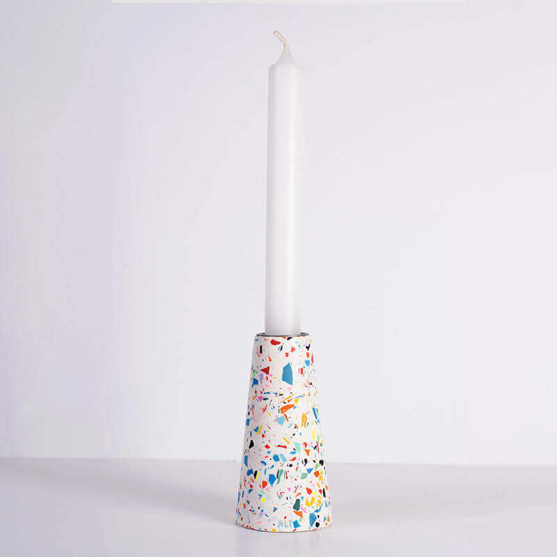 A tall white taper candle standing upright in a white and rainbow terrazzo patterned taper candle holder with a silver-toned top, shown on a white background
