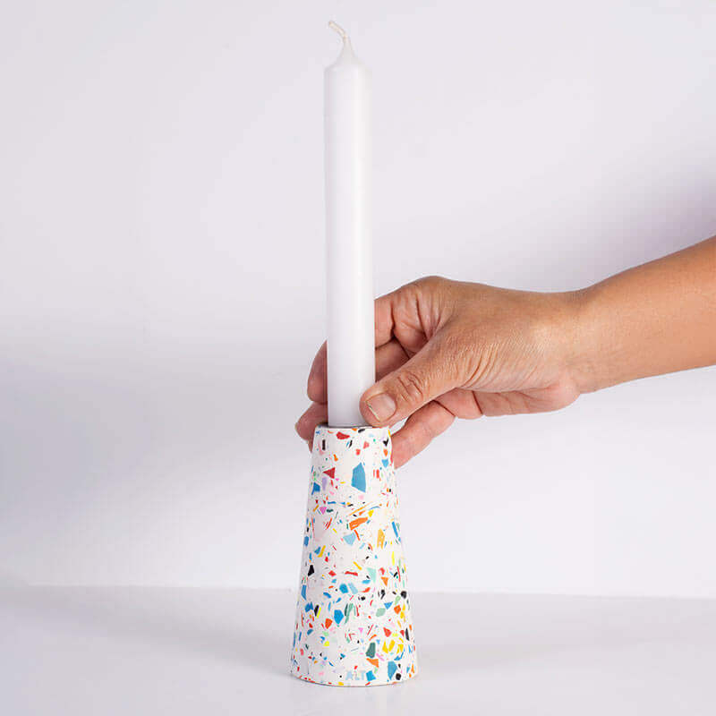 A hand holding a white taper candle in a white and rainbow terrazzo patterned taper candle holder with a silver-toned top, shown on a white background
