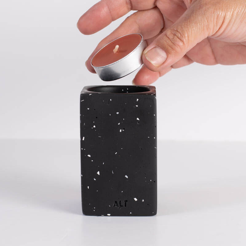 Hand placing a candle into a black speckled container with 'ALT' branding on a white background